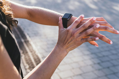 From above crop anonymous athletic female runner checking pulse on fitness tracker while standing on paved pathway during training in city on sunny day