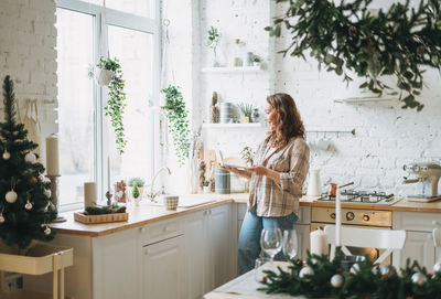 Attractive smiling woman with curly hair in plaid shirt with laptop near window at bright kitchen