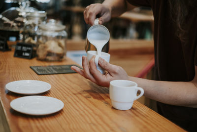 Midsection of woman pouring milk in coffee on wooden table in cafe