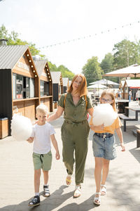 Full body of mother with children eating sweet cotton candy while walking together on fairground on sunny summer day in city