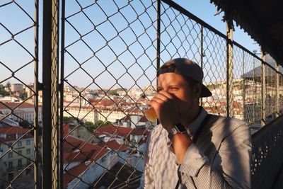Man drinking by chainlink fence against sky