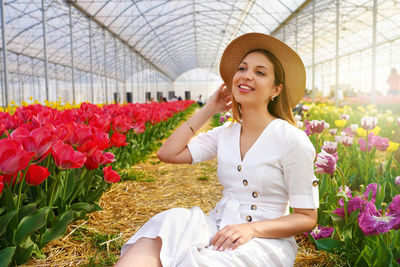 Pretty beautiful girl with straw hat sitting between tulips field in greenhouse