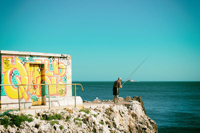 Rear view of man fishing in sea against clear blue sky