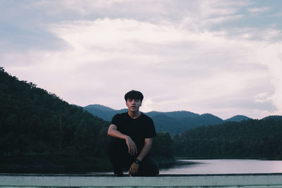 Portrait of young man sitting on mountain against sky