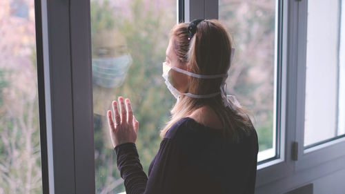 Woman wearing mask looking through window at home