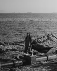 Full length of young woman standing on concrete over rock formation against sea