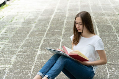Teenage girl writing in paper with book while sitting on footpath