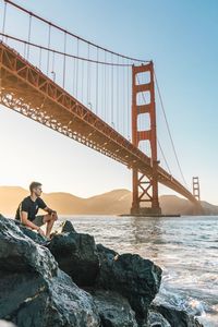 Young man sitting on rock by sea under golden gate bridge against clear sky