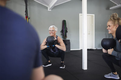 Smiling women exercising with kettlebells while squatting in gym