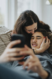 Teenage boy using mobile phone while mother kissing him in living room