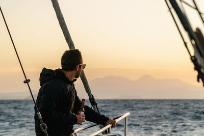 Male on boat bow looking at horizon with beer in hand during sunset at sea on a sailboat.
