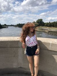 Beautiful young woman standing by retaining wall against sky