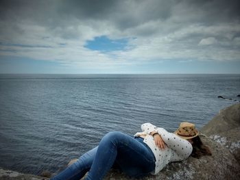 Woman with face covered by hat relaxing on rock against sea