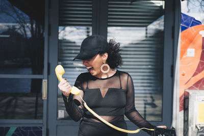 Angry young woman holding telephone receiver connected with mobile phone while standing against building