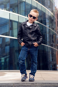 Baby boy in sunglasses and black leather jacket smiling on the background of the glass building