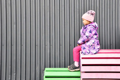 Side view of girl in warm clothing looking away while sitting on bench against wall