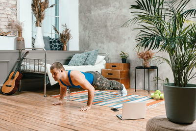 Cheerful sportsman with black hair does push-ups and watching online workout from laptop in bedroom