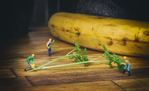 Close-up of construction worker figurines cutting cilantro by banana on wooden table