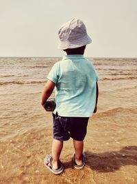 Rear view of boy standing at beach against clear sky