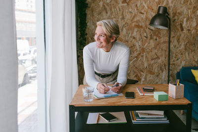 Focused adult businesswoman looking away while taking notes of plan on clipboard bending on wooden table in light office