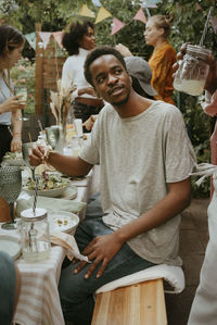 Young man eating salad and talking with friend while sitting on bench at dinner party in back yard