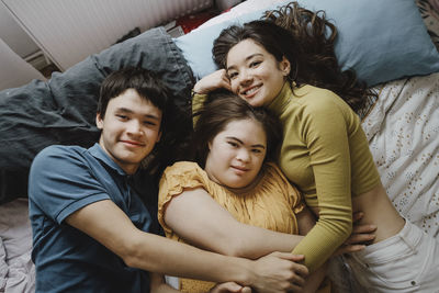 Directly above portrait of siblings embracing down syndrome sister while lying down on bed at home