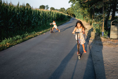 Two children riding red scooters on a country road next to corn field