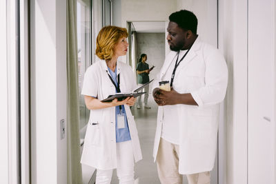 Senior female doctor discussing over medical record with young male colleague while walking in hospital corridor