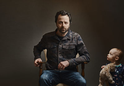 Portrait of father sitting on chair with daughter against gray background