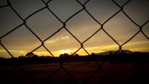 Silhouette landscape seen through chainlink fence against sky during sunset