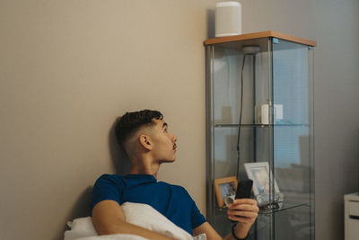 Boy with mobile phone looking away while sitting against wall at home