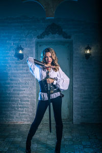 Portrait of woman in costume holding sword standing against door of abandoned building at night