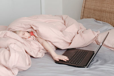 Sleepy tired girl lying in bed under blanket and learning in virtual online school class. 