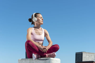 Attractive fit woman sitting crossed legged slistening to music with headphones outdoors in the sun.