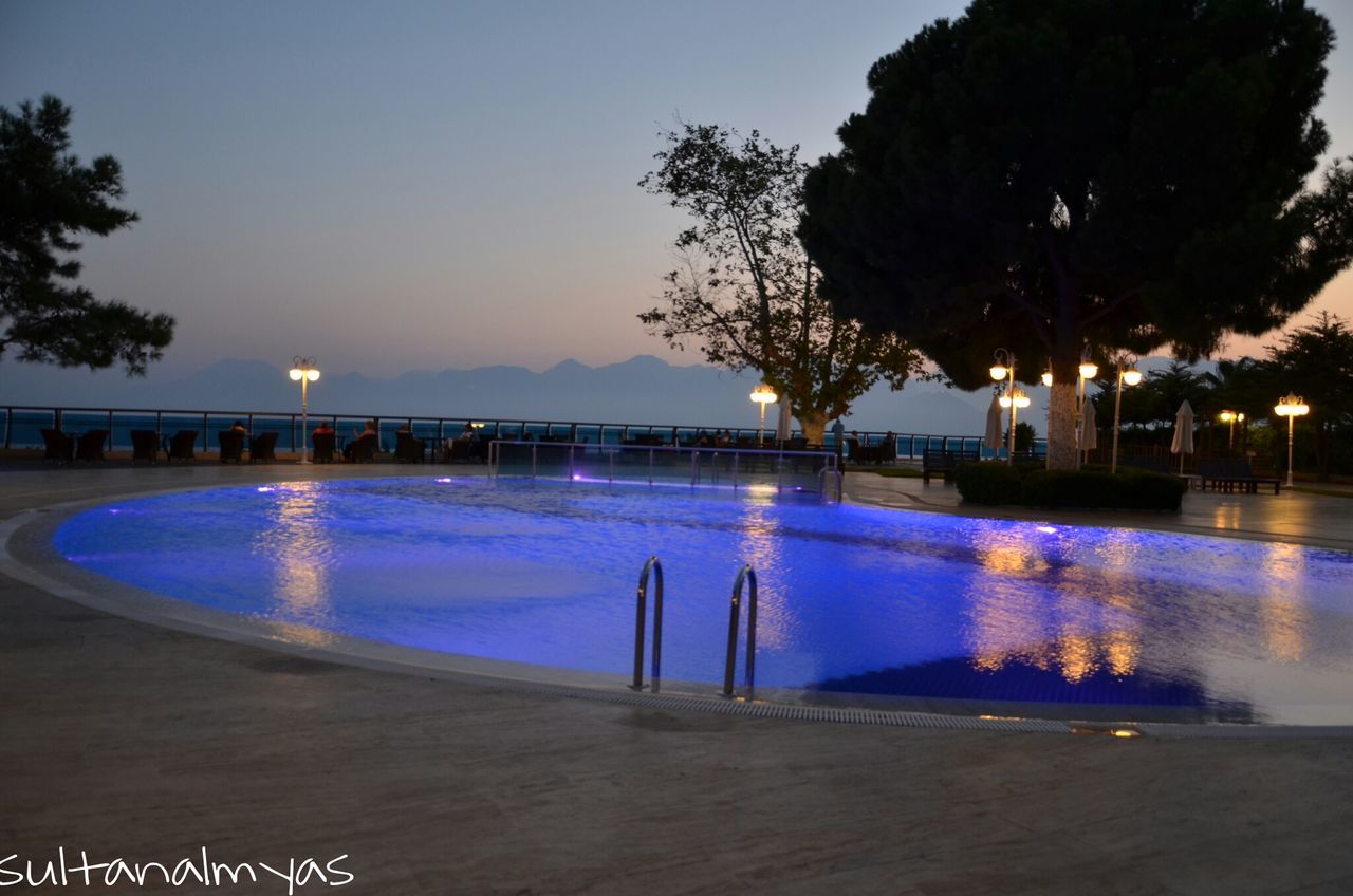 water, illuminated, tree, swimming pool, blue, sea, clear sky, reflection, sky, lighting equipment, scenics, tranquility, tranquil scene, night, street light, nature, beach, outdoors, dusk, built structure