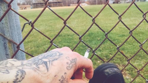 Cropped hand of man holding cigarette against fence