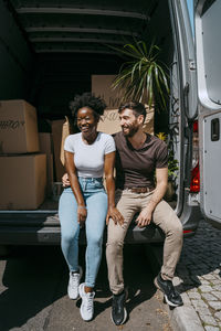 Happy multiracial couple sitting together in van trunk full of boxes