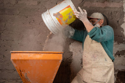 Focused male worker in apron and respirator pouring dry mix from bucket into metal plaster station while working in ceramic workshop