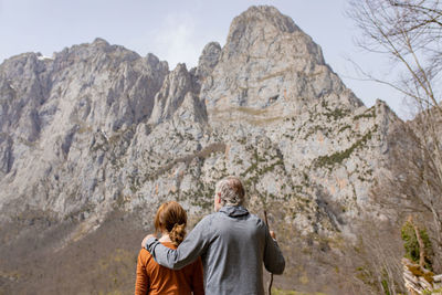 Back view of unrecognizable gray haired man with stick embracing woman while standing in front of high rocky picos de europa mountain range and admiring landscape as hiking together in castile and leon in spain