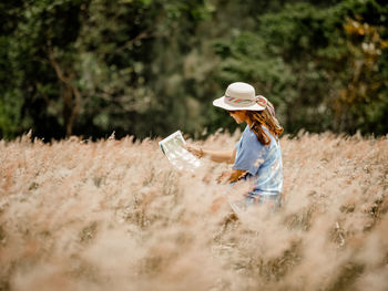 Woman wearing hat while reading map on field against trees