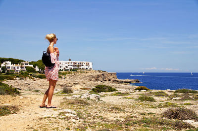 Side view of woman looking at sea while standing footpath against blue sky