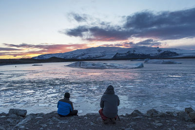 Rear view of people sitting at frozen lake against sky during sunset