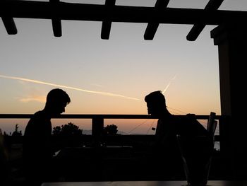 Silhouette people sitting by railing against sky during sunset