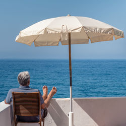 Rear view of man looking at sea while sitting on chair by parasol at building terrace 
