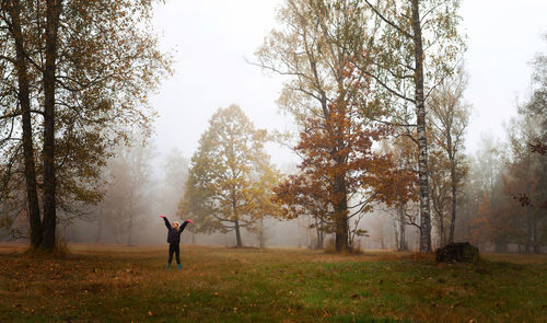 Girl standing with arms raised on field at forest during foggy weather