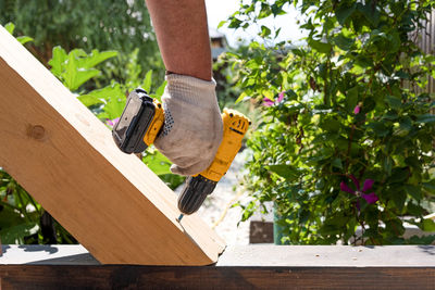 A carpenter works with a yellow electric screwdriver, twists a screw into a wooden plank beam