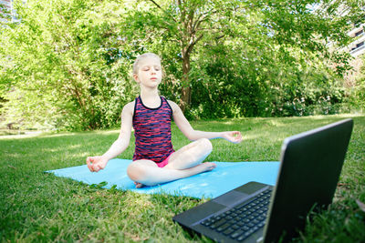 Full length of girl sitting meditating on grass by laptop