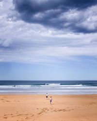 Rear view of shirtless men running at beach against cloudy sky