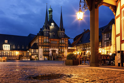 Illuminated street amidst buildings against sky at dusk