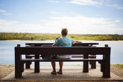 Rear view of senior woman using laptop while sitting on bench at lakeshore against sky